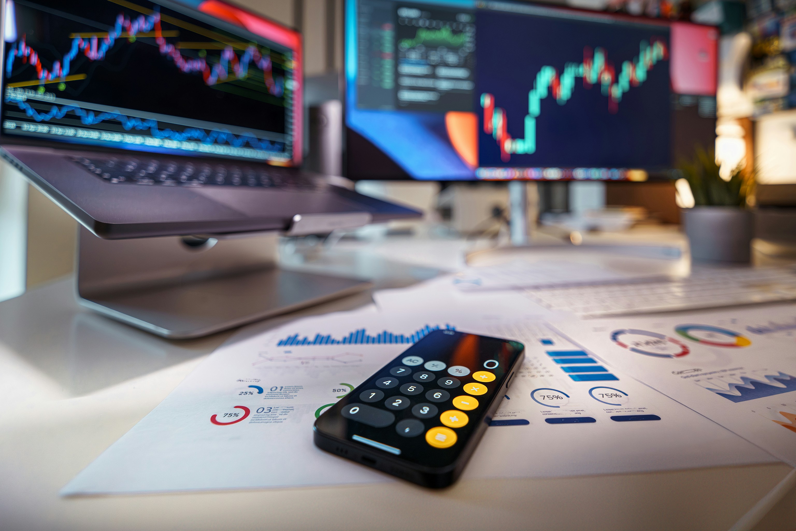 A trading desk with printed charts and statistics, a calculator app open on a mobile phone, and two computer screens displaying blurred candlestick charts — representing the data-intensive infrastructure environment of stockbrokers and proprietary trading firms.